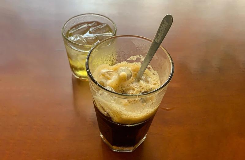 Ly c&agrave; ph&ecirc; sữa đ&aacute; v&agrave; ly tr&agrave; đ&aacute; tr&ecirc;n b&agrave;n gỗ, c&oacute; th&igrave;a trong ly c&agrave; ph&ecirc;.