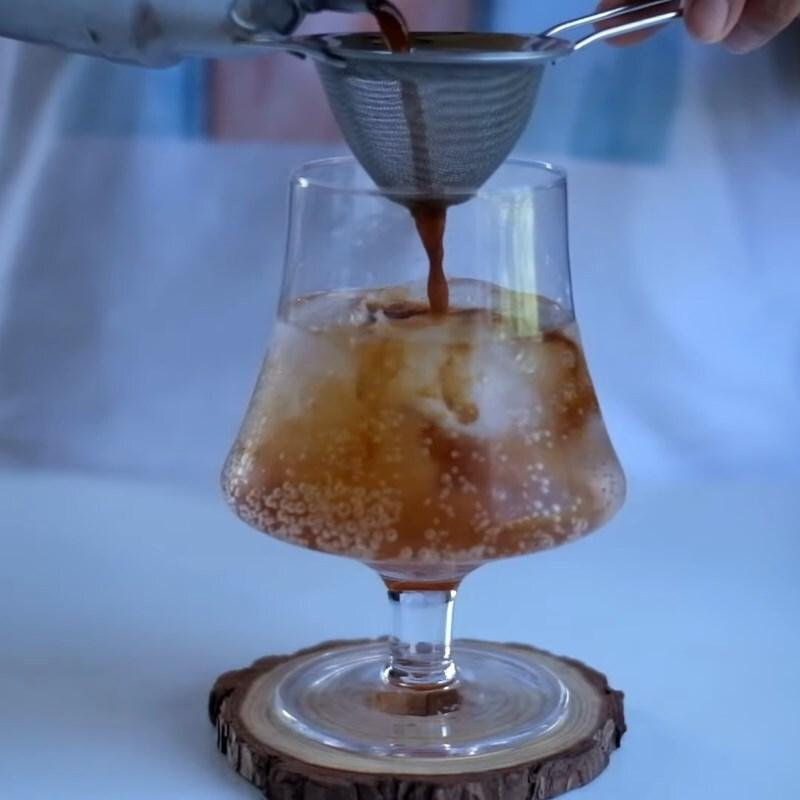 Đồ uống m&agrave;u n&acirc;u được r&oacute;t qua r&acirc;y lọc v&agrave;o ly thủy tinh c&oacute; đ&aacute;, đặt tr&ecirc;n đế gỗ.