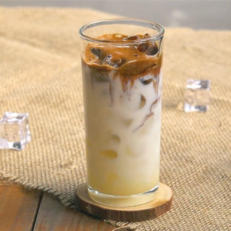 Ly c&agrave; ph&ecirc; sữa đ&aacute; tr&ecirc;n đế gỗ, c&oacute; lớp bọt v&agrave; đ&aacute; vi&ecirc;n, đặt tr&ecirc;n nền vải m&agrave;u n&acirc;u nhạt.