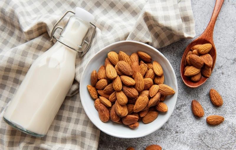 B&igrave;nh sữa hạnh nh&acirc;n b&ecirc;n cạnh b&aacute;t v&agrave; muỗng chứa hạt hạnh nh&acirc;n tr&ecirc;n khăn kẻ &ocirc; m&agrave;u x&aacute;m.