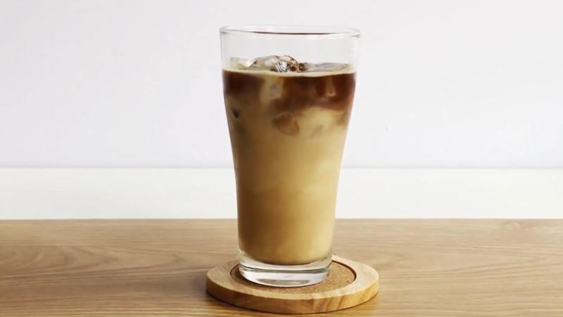 Ly c&agrave; ph&ecirc; sữa đ&aacute; tr&ecirc;n b&agrave;n gỗ, đặt tr&ecirc;n đế l&oacute;t ly tr&ograve;n, với nền trắng ph&iacute;a sau.