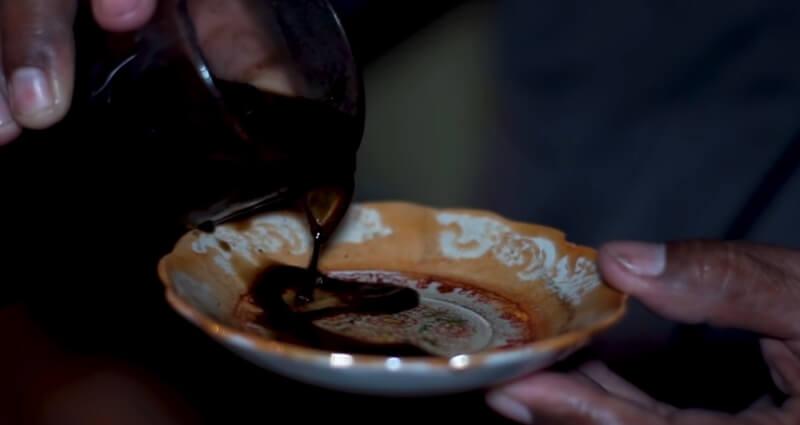 Người đang r&oacute;t chất lỏng m&agrave;u đen từ cốc v&agrave;o đĩa nhỏ hoa văn, cầm đĩa bằng tay phải.