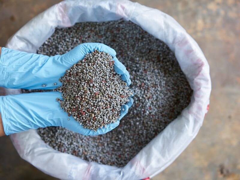 Đôi tay đeo găng đang cầm hạt phân bón từ bao lớn, bề mặt sàn phía dưới.