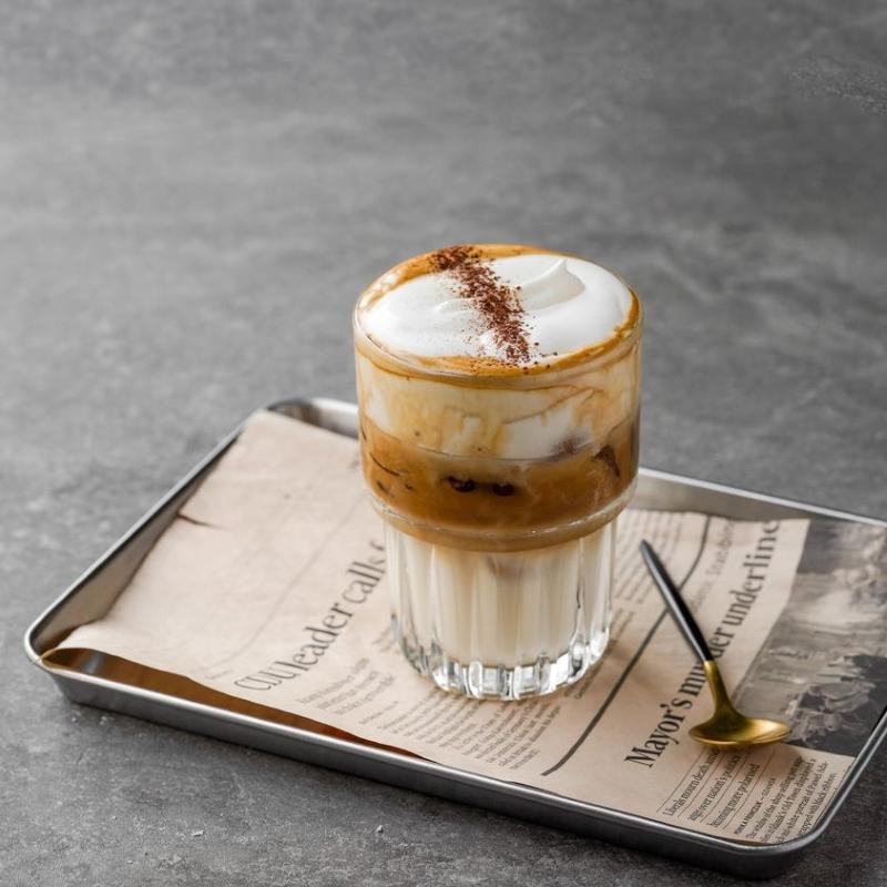 Ly c&agrave; ph&ecirc; sữa đ&aacute; với lớp bọt sữa, đặt tr&ecirc;n khay c&ugrave;ng th&igrave;a v&agrave; tờ b&aacute;o tr&ecirc;n b&agrave;n x&aacute;m.