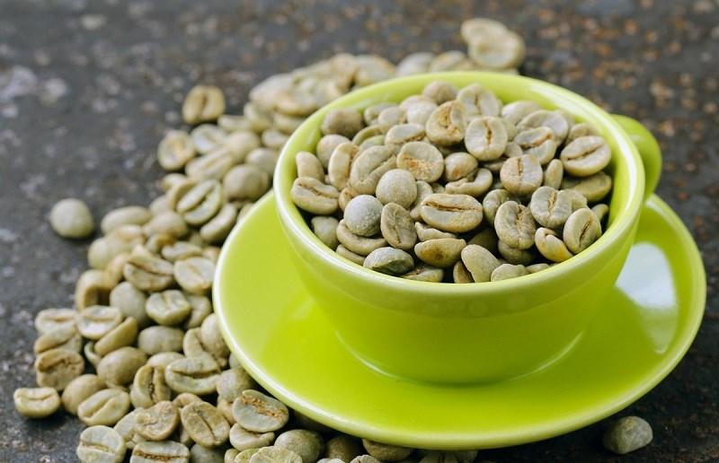 Tách màu xanh chứa hạt cà phê xanh, xung quanh là nhiều hạt cà phê xanh nằm trên bề mặt tối.