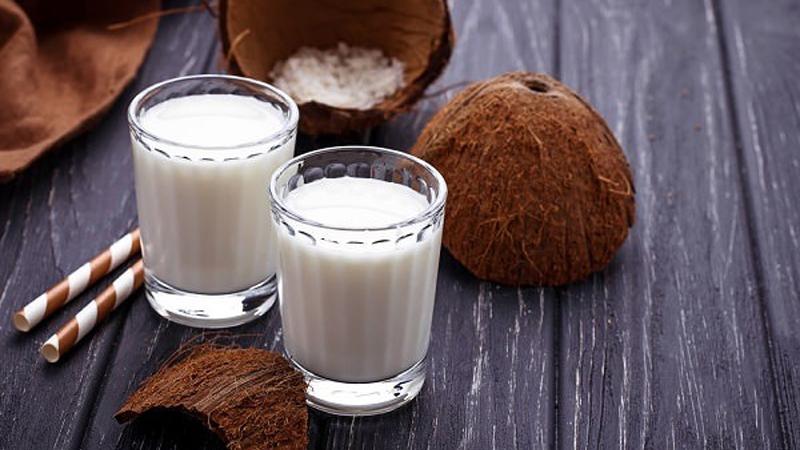 Hai ly sữa dừa trên bàn gỗ, bên cạnh là trái dừa và ống hút sọc.