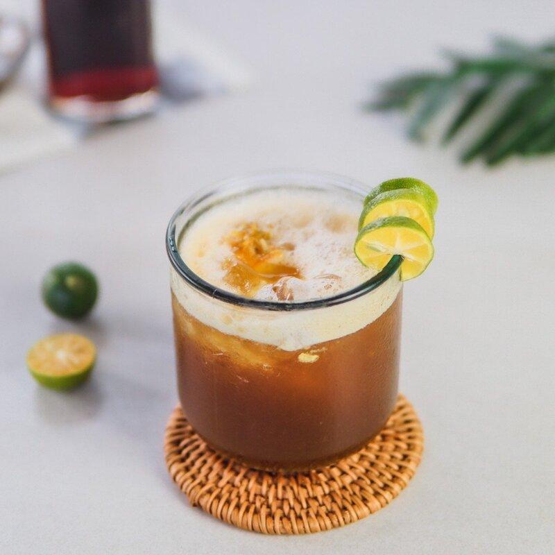 Ly cà phê đá với lát chanh trang trí, đặt trên đế lót bằng mây, bên cạnh là vài quả chanh.