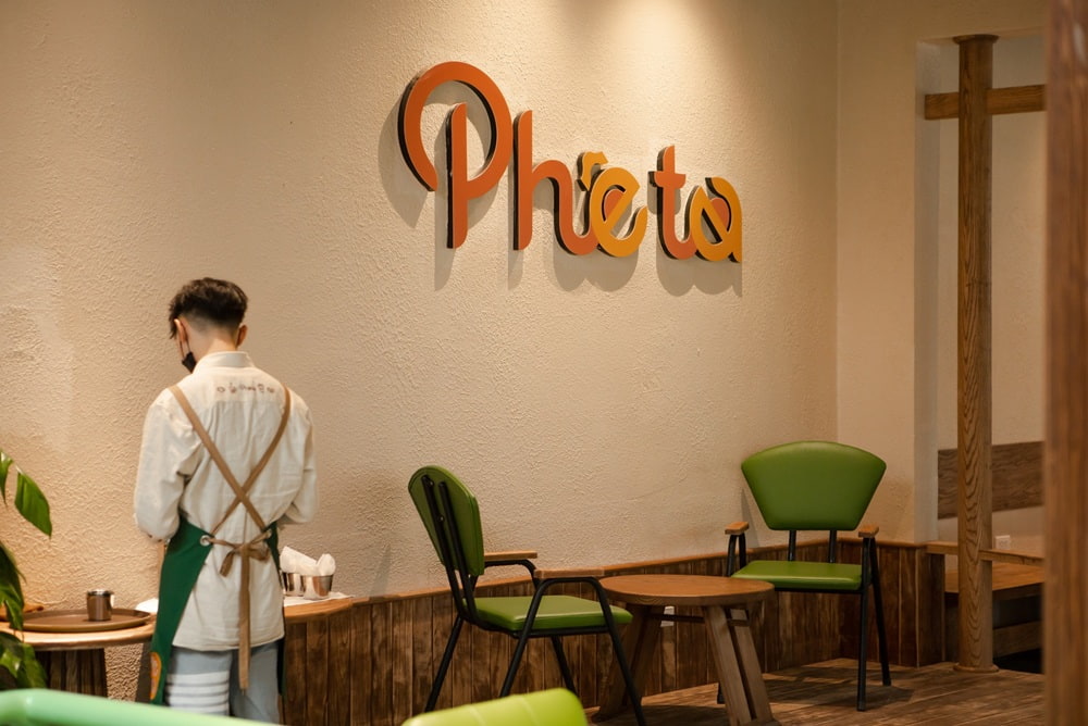 Khám Phá 15+ Quán Cafe View Đẹp, Đồ Uống Ngon Tại Hoàng Cầu 19 Nhân viên đứng cạnh bàn trong quán cà phê với bảng hiệu "Phê Tê" trên tường, ghế màu xanh lá.