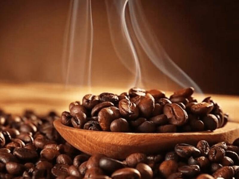 cac-loai-ca-phe-robusta-3 Hạt cà phê rang bốc khói trên muỗng gỗ, nền mờ với tông màu nâu ấm áp.