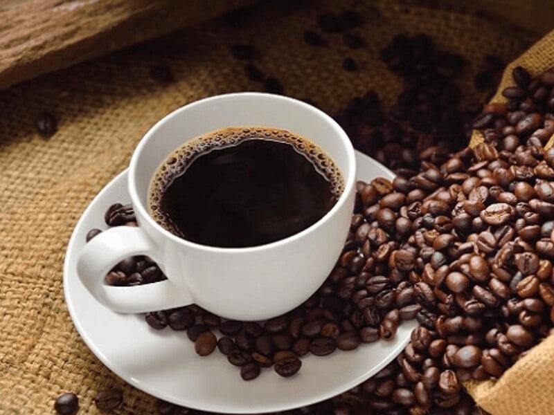 cac-loai-ca-phe-robusta-4 Tách cà phê đen trên đĩa trắng, xung quanh là hạt cà phê, đặt trên nền vải bố màu nâu.