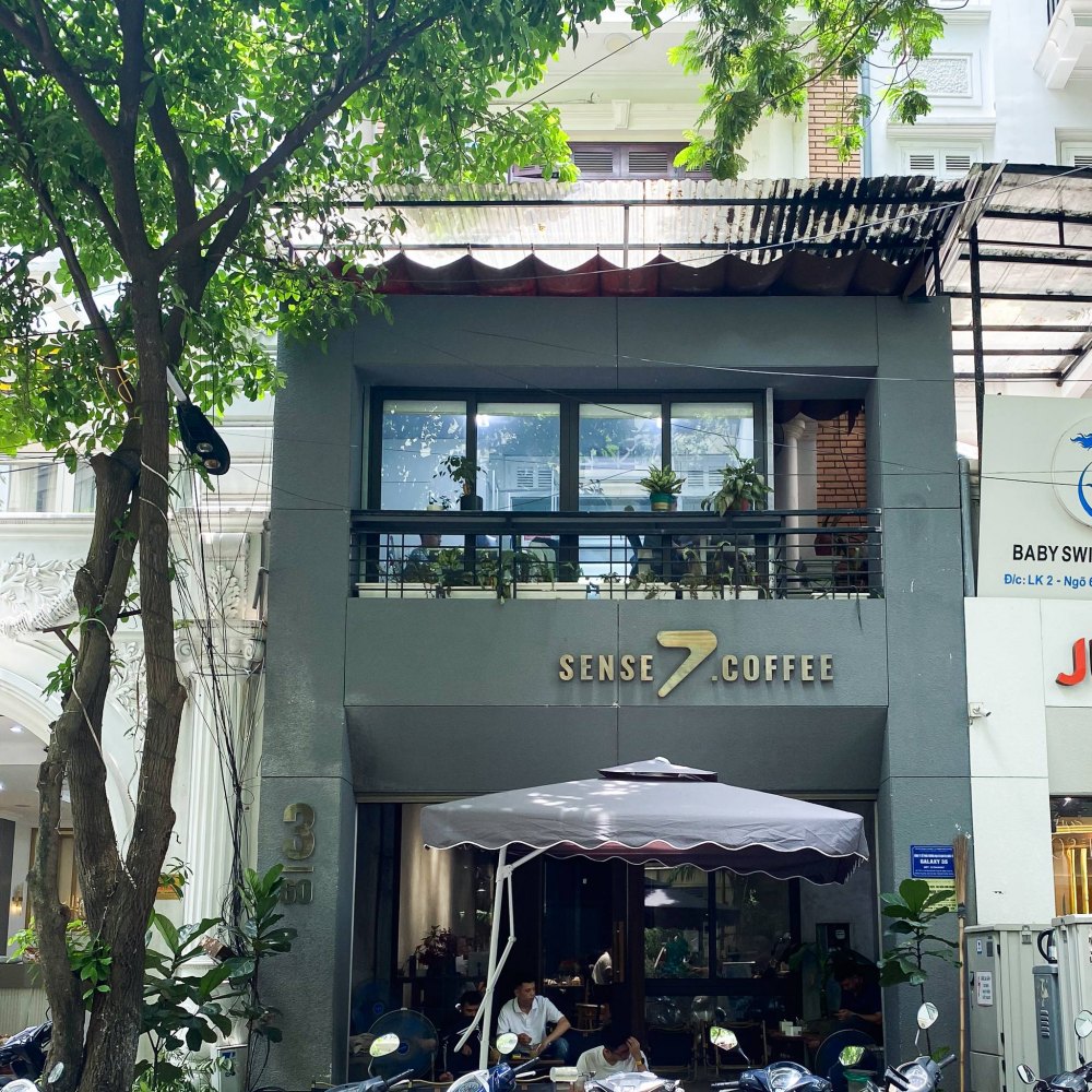 Mặt tiền một quán cà phê với cây xanh phía trước, có biển hiệu "Sense 7 Coffee" và một ô che nắng.