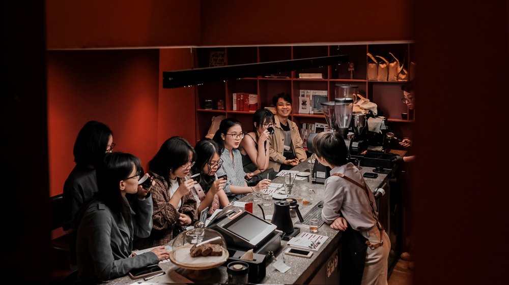Một nhóm người ngồi tại quầy bar trong quán cà phê, trò chuyện và thưởng thức đồ uống.