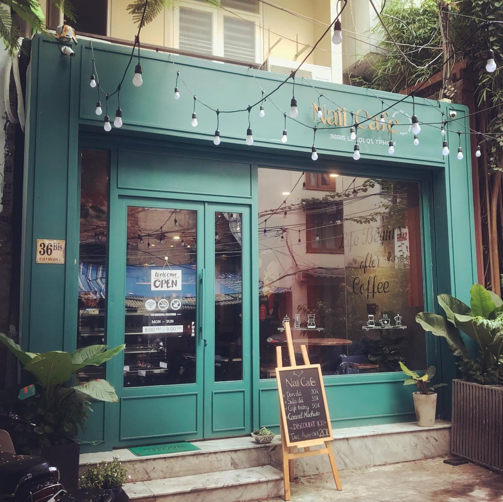 Mặt tiền quán cà phê xanh với bảng hiệu và bảng đen ghi món uống, có dây đèn trang trí phía trên.