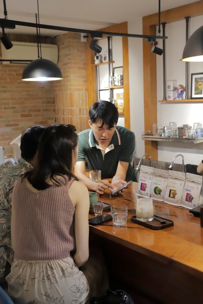 Nhóm người ngồi quanh quầy trong quán cà phê, một người đang chỉ vào màn hình điện thoại.