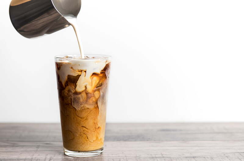 cach-lam-bac-xiu-4-tang-4 Ly cà phê đá đang được rót sữa vào từ một chiếc bình kim loại trên nền trắng.