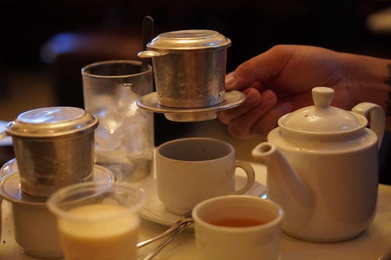 cach-lam-bac-xiu-4-tang-5 Một bàn với bộ pha cà phê phin, ấm trà trắng, cốc nước và cốc sữa, tay người đang cầm phin cà phê.