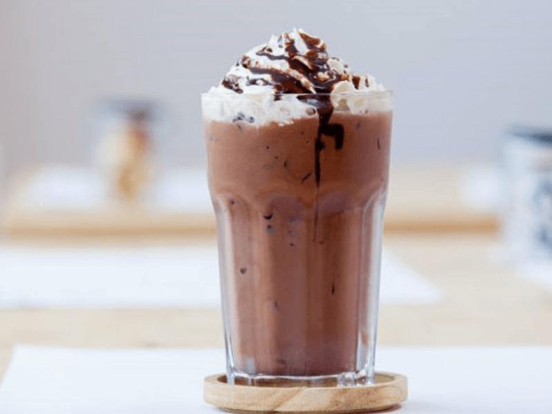 Ly chocolate đá xay với kem tươi và sốt chocolate, đặt trên bàn cùng đế lót ly gỗ.