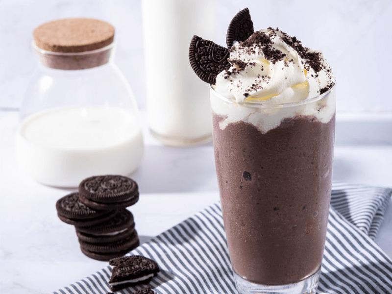 Ly sinh tố chocolate với kem tươi, bánh quy vụn trang trí. Bình sữa và bánh quy bên cạnh trên bàn vải sọc.