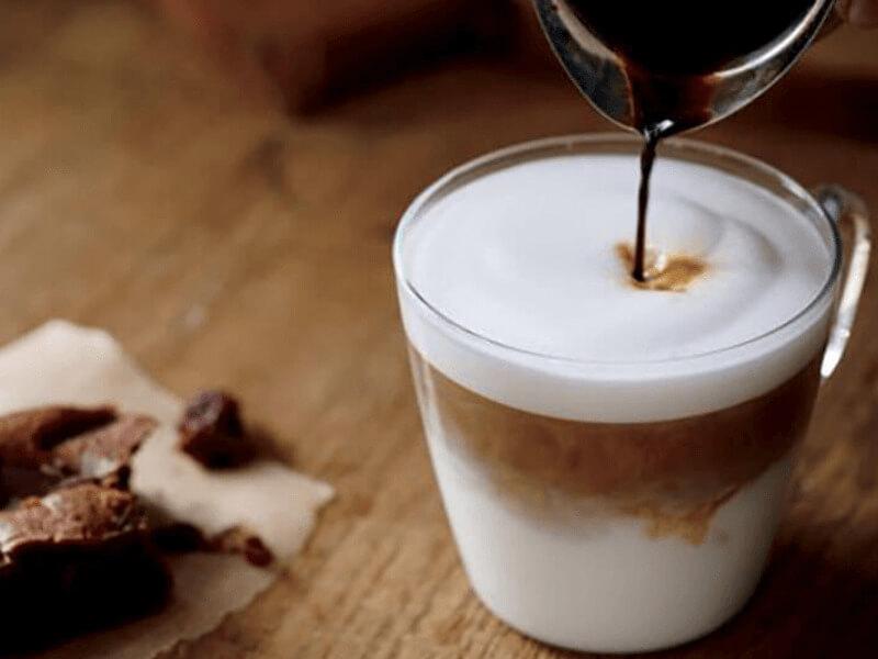 Ly cà phê sữa với lớp bọt mịn, đang được rót thêm cà phê từ một chiếc cốc nhỏ trên nền gỗ.