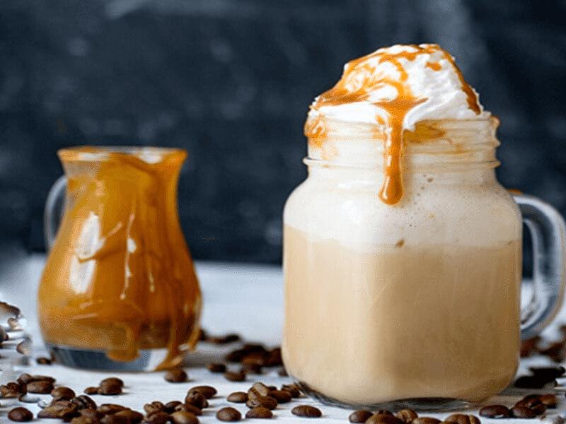 Ly cà phê kem với caramel, bên cạnh là lọ nhỏ đựng caramel, trên nền nhiều hạt cà phê.