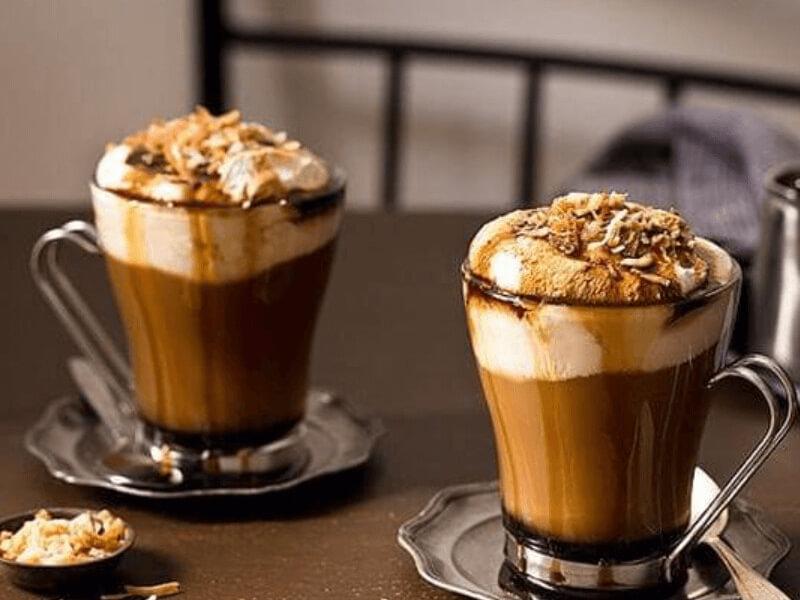 Hai ly cà phê với kem tươi và topping hạt dẻ, đặt trên bàn với đĩa lót sắt.