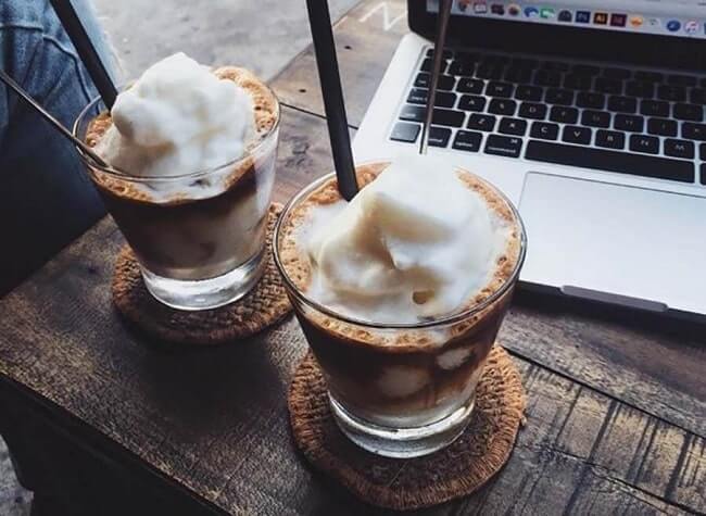 Hai ly cà phê sữa đá với lớp bọt trắng, đặt trên bàn gỗ cạnh máy tính xách tay.