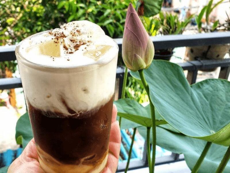 Ly cà phê sữa đá với bọt kem, bên cạnh là một bông hoa sen chưa nở, nền là cây xanh và ban công.