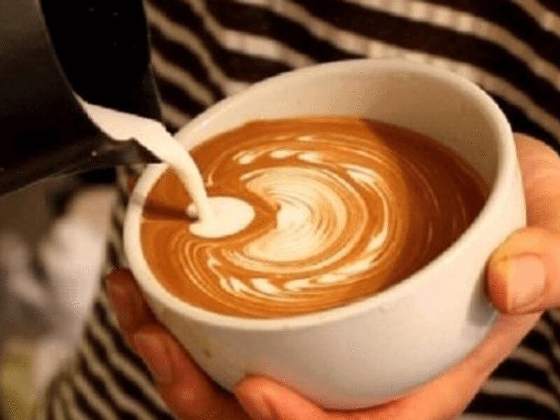 Tay đang rót sữa vào cốc cà phê, tạo hình xoáy nghệ thuật trên bề mặt.