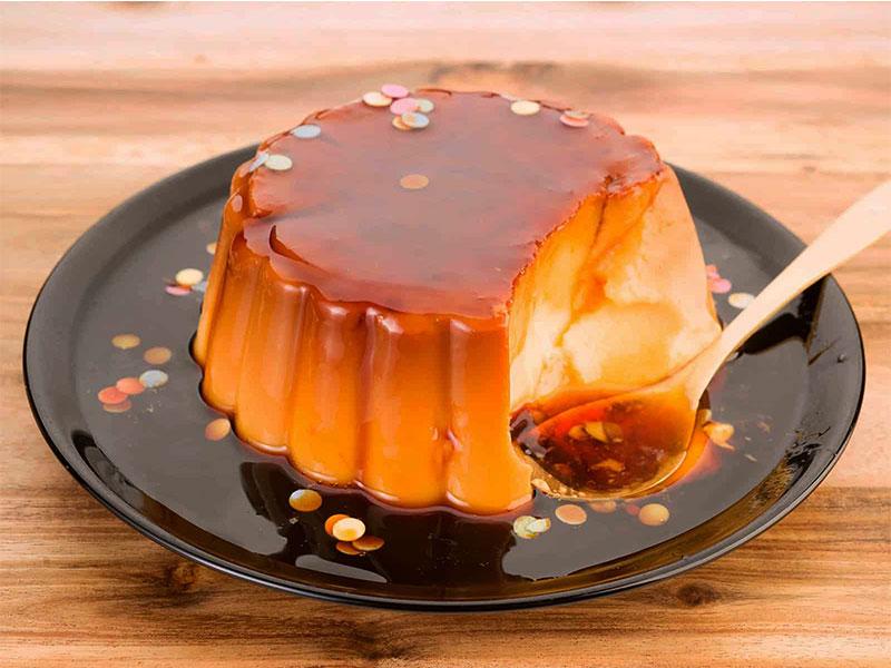 cach-lam-sot-ca-phe-an-banh-flan-2 Bánh flan caramel trên đĩa đen, có rắc vụn màu, một phần bị ăn dở, bên cạnh là thìa gỗ.