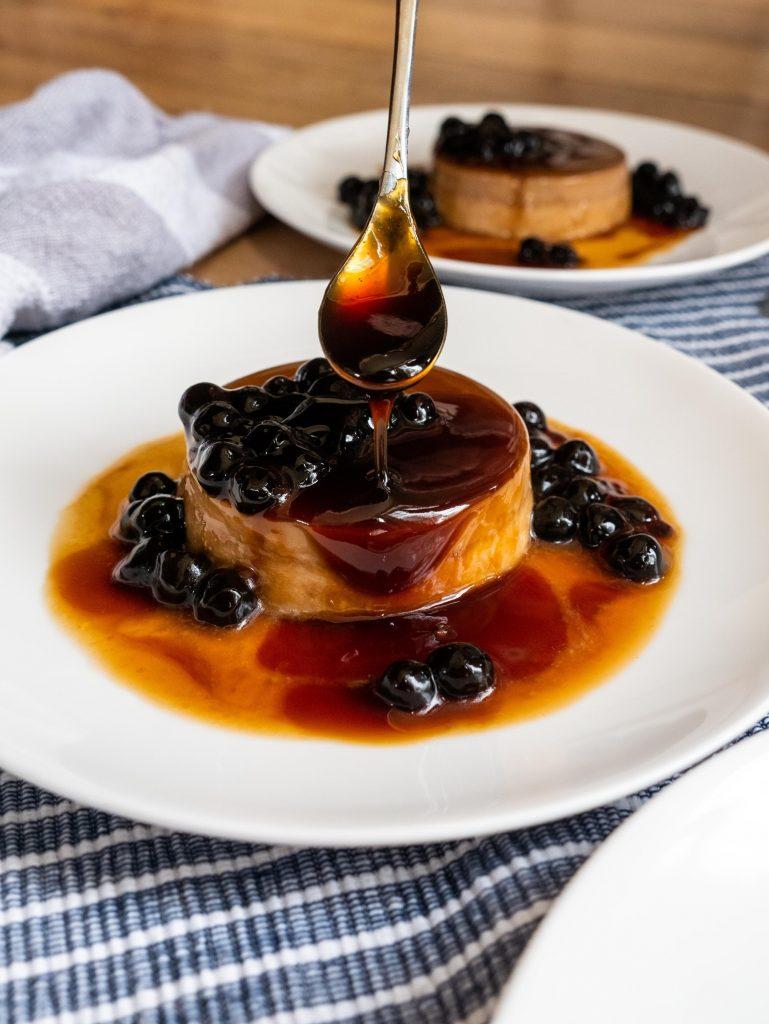 cach-lam-sot-ca-phe-an-banh-flan-5 Bánh flan trên đĩa trắng, phủ sốt caramel và trân châu, muỗng đang rót sốt lên bánh, khăn trải bàn sọc xanh.