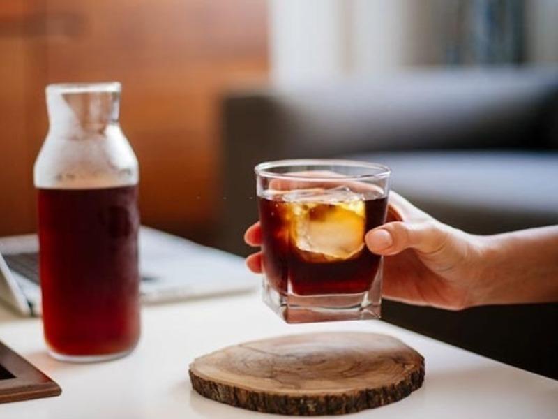 Một tay cầm ly nước màu nâu đá, bên cạnh là chai thủy tinh đựng cùng loại nước, đặt trên bàn trắng.