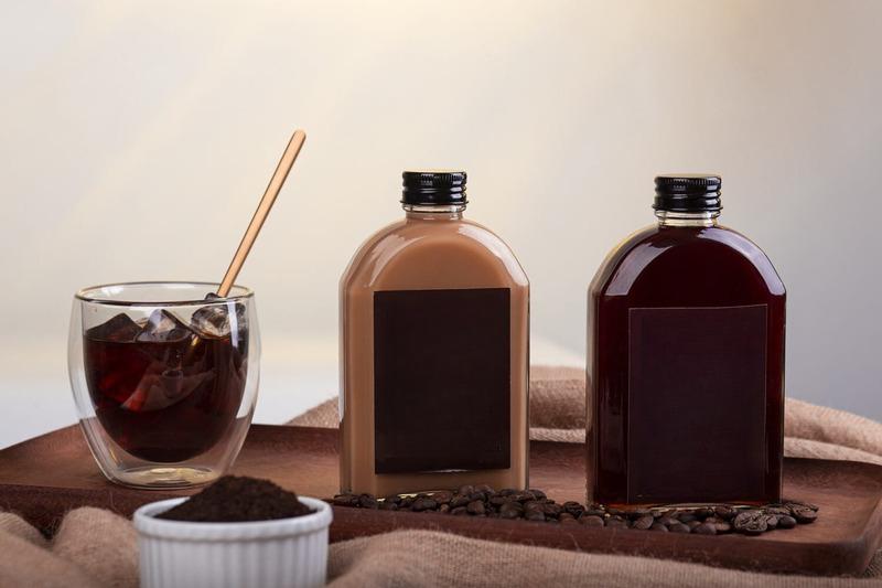 cach-pha-ca-phe-dong-chai-5 Hai chai nước tối màu và một ly cà phê đá trên bàn, xung quanh có hạt cà phê và bột cà phê.