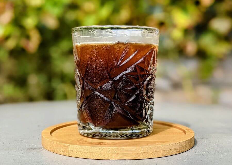 Ly cà phê đá trong ly thủy tinh có hoa văn, đặt trên đĩa gỗ, nền mờ xanh lá.