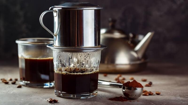 Ly cà phê phin đang pha trên bàn, bên cạnh là ấm nước và cà phê xay.