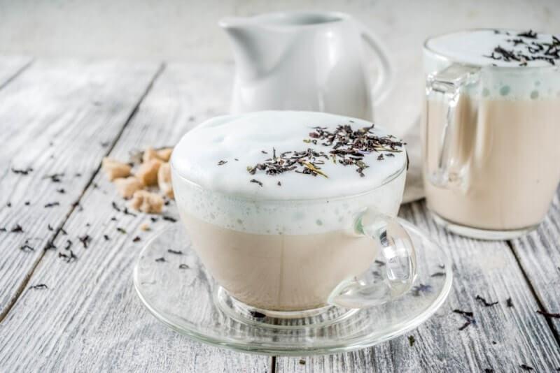 Hai ly trà sữa với bọt sữa và lá trà khô trên mặt, đặt trên bàn gỗ trắng cùng bình sữa nhỏ phía sau.
