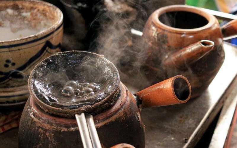 Ấm đất đang đun với nước s&ocirc;i bốc hơi, b&ecirc;n cạnh l&agrave; một ấm kh&aacute;c v&agrave; một b&aacute;t sứ c&oacute; hoa văn.