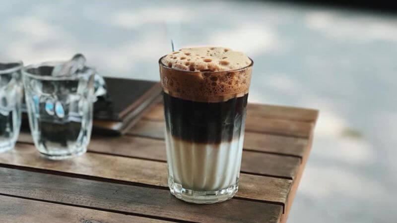 Ly cà phê sữa đá với lớp bọt trên cùng đặt trên bàn gỗ ngoài trời, cạnh là hai ly nước.