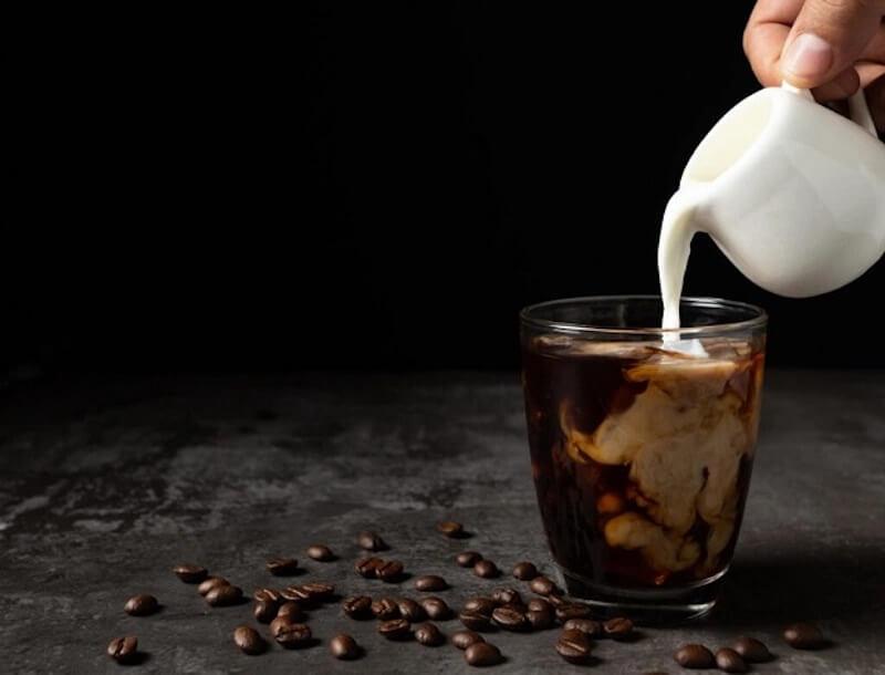 Tay rót sữa vào ly cà phê đen trên nền tối, hạt cà phê rải trên bàn.
