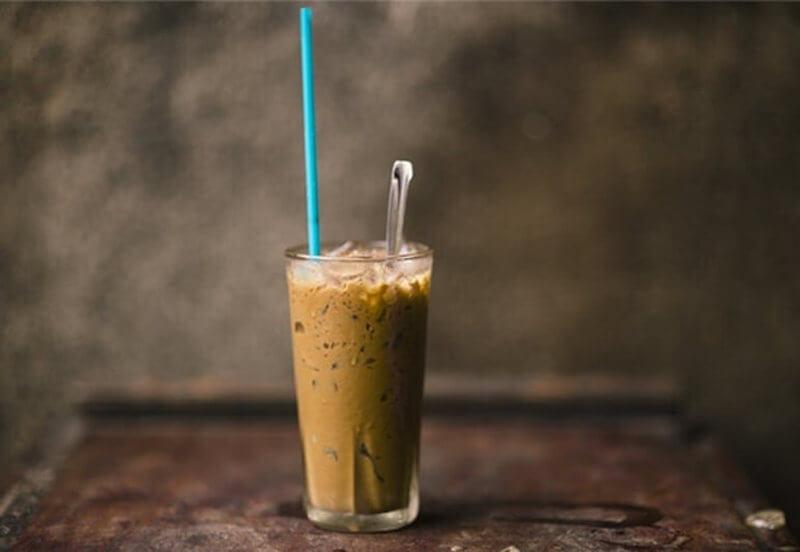 Ly cà phê sữa đá trên bàn, có ống hút xanh và muỗng, nền tường mờ.