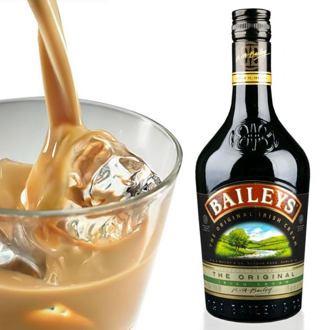 Chai rượu sữa Baileys cạnh ly chứa đồ uống màu nâu đổ vào đá.