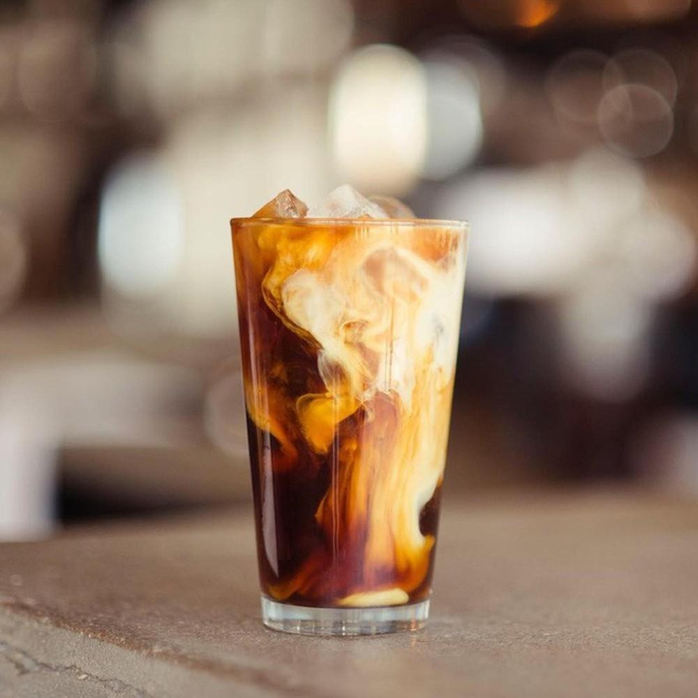 Ly cà phê đá với sữa đang hòa tan, đặt trên bàn, nền mờ.