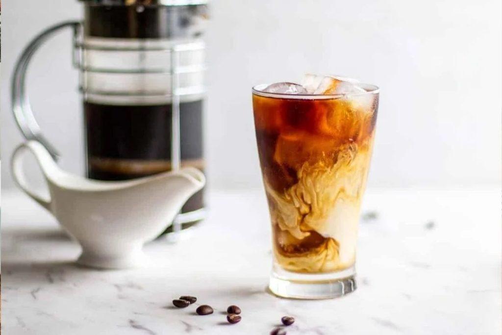 Ly cà phê sữa đá với lớp kem hòa quyện, bình cà phê và hũ đựng sữa trên nền bàn đá trắng.