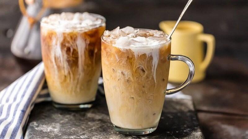 Hai ly cà phê sữa đá trên bàn, có ống hút và thìa khuấy, với một bình màu vàng ở phía sau.