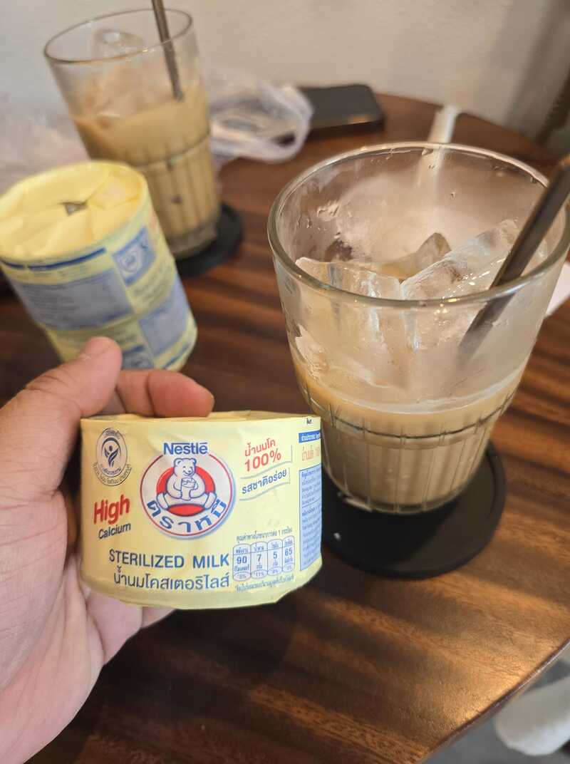 Người cầm hộp sữa tiệt tr&ugrave;ng, b&ecirc;n cạnh l&agrave; ly c&agrave; ph&ecirc; sữa đ&aacute; tr&ecirc;n b&agrave;n gỗ.