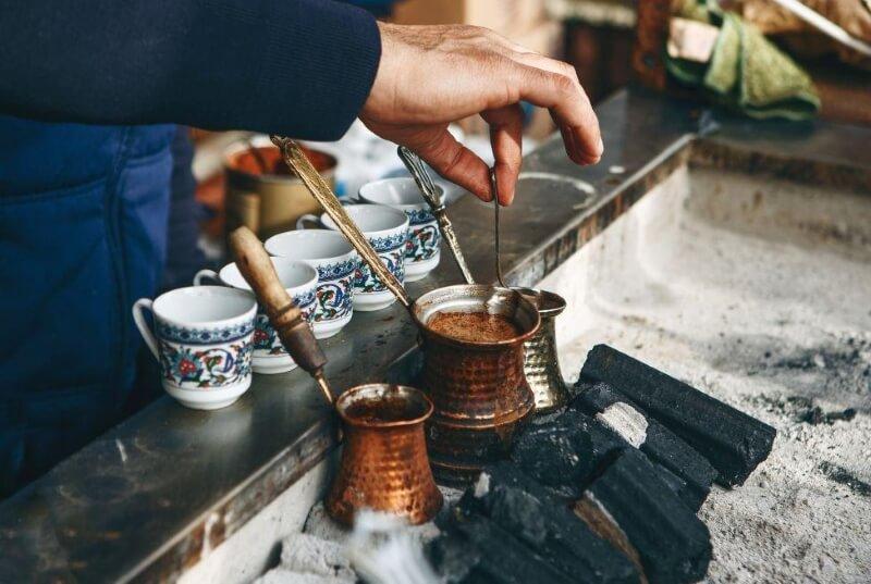 Người pha cà phê Thổ Nhĩ Kỳ truyền thống trên bếp than, bên cạnh là nhiều tách cà phê trang trí hoa văn.