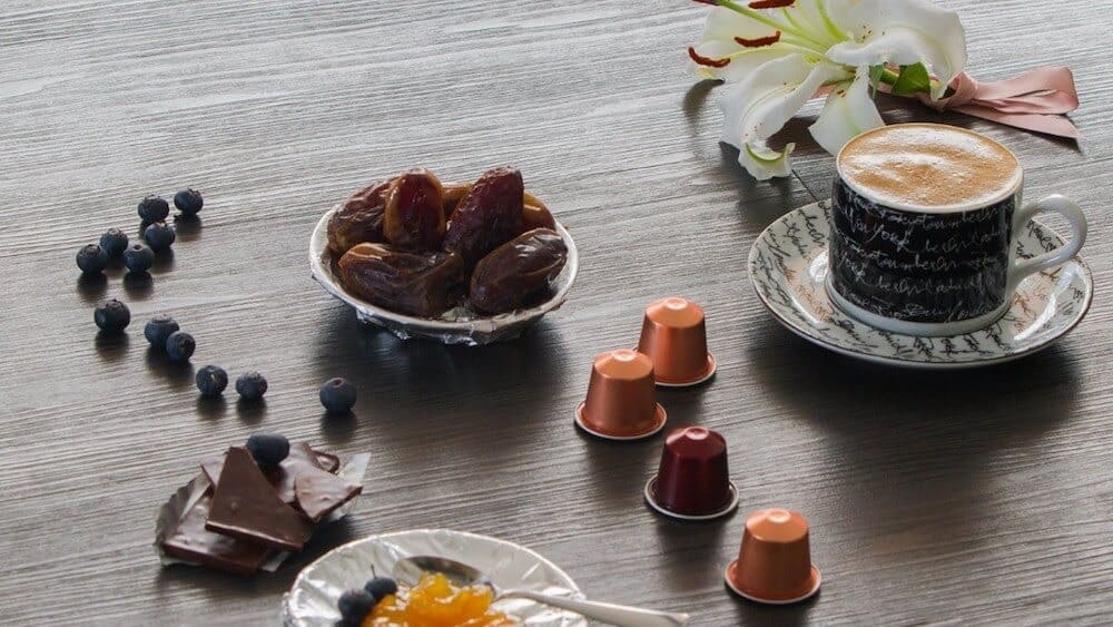 cach-pha-ca-phe-vien-nen-khong-dung-may-1 Cốc cà phê trên bàn gỗ cùng quả chà là, việt quất, sô cô la, viên nén cà phê và hoa trang trí.