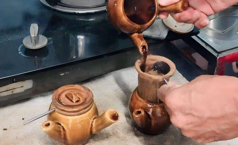 Người đang đổ cà phê từ phin vào một ấm gốm trên bếp, cạnh đó là một ấm trà nhỏ khác.