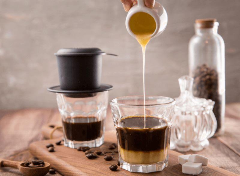 Ly cà phê phin với sữa đặc đang được rót vào, đặt trên bàn gỗ cùng nhiều vật dụng pha chế khác.