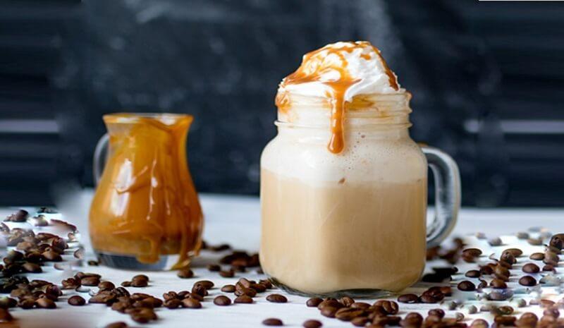 Ly cà phê sữa đá có kem tươi và caramel, cạnh đó là lọ caramel, xung quanh có hạt cà phê.
