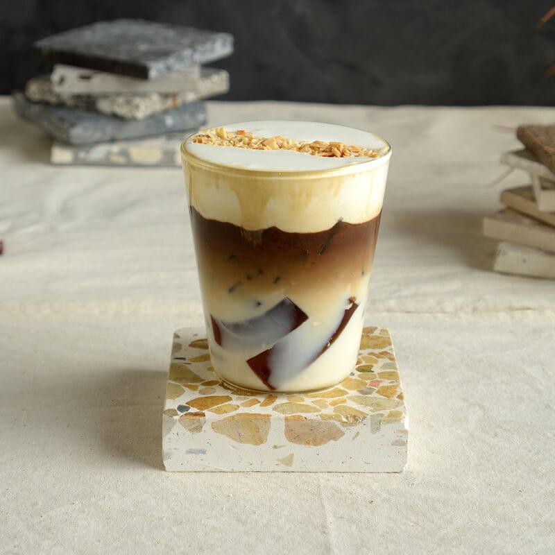 Ly cà phê sữa đá với lớp kem sánh mịn và topping hạnh nhân, đặt trên bàn cùng đế lót đá trang trí.